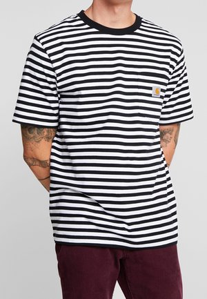 Mand iført sort-hvidt vandret striped t-shirt med lille lomme og bordeaux bukser, tatoveringer synlige på begge underarme.