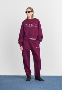 Persona che indossa un completo sportivo Nike viola e sneakers nere, in piedi su un tappeto blu contro un muro bianco, con occhiali da sole neri.
