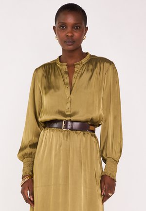 Femme portant une robe longue en satin olive à manches longues avec des poignets froncés, une ceinture marron foncé, des boucles d'oreilles créoles dorées et des bracelets dorés à un poignet.