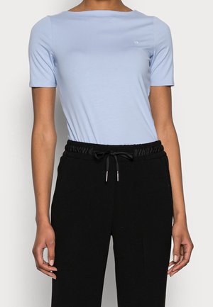 Osoba ubrana w jasnoniebieską bluzkę z krótkim rękawem i dekoltem typu łódka oraz czarne spodnie ze sznurkiem na jednolitym tle.