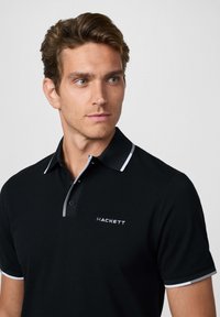 Camiseta polo negra con una tapeta de dos botones, cuello ajustado con ribetes en blanco y gris, y un logo de "HACKETT" en el lado izquierdo del pecho.