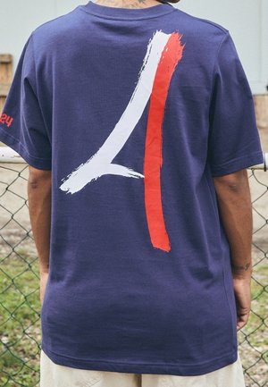 Person trägt ein dunkelblaues T-Shirt mit einem großen weißen und roten Pinselstrich-Design auf dem Rücken und steht vor einem Maschendrahtzaun.