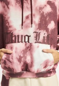 Felpa tie-dye rosa e bianca con tasca a marsupio frontale e testo nero "Thug Life", dotata di coulisse con punte metalliche.