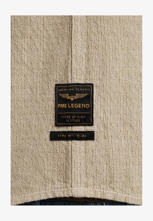 Beige Textil mit strukturierter Oberfläche, auf dem ein schwarzes Etikett mit der Aufschrift "PME LEGEND" und Informationen über Höhentests sowie Typennummer zu sehen ist.