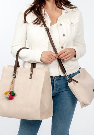 Femme portant une veste blanche et un jean bleu, tenant un sac fourre-tout beige en tissu tressé avec des pompons colorés et une pochette assortie à bandoulière marron.