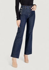 Jeans flare in denim scuro con vita alta, dotati di bottoni dorati laterali e cuciture a contrasto. Texture liscia e silhouette aderente.