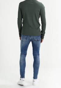 Vihreä ribatattu pitkähihaisena paita, siniset slim-fit farkut, valkoiset tennarit mustalla logolla. Farkuissa näkyvät saumat, yksinkertainen muotoilu.