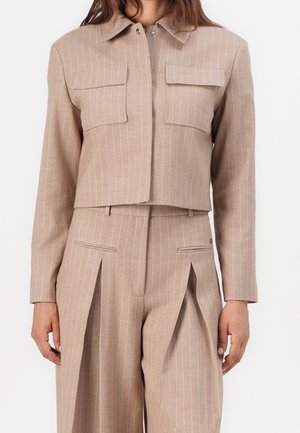 Vrouw draagt een beige cropped jasje met krijtstreepjes en klepzakken en bijpassende hoog getailleerde wijde broek met voorvouwen.