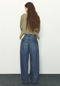 Žena s dlhými hnedými vlasmi, oblečená v žltom a hnedom pruhovanom svetri a širokých modrých džínsoch, stojaca pri bielej stene.