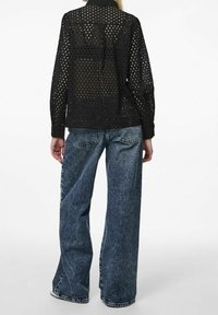 Černá poloprůhledná košile s perforovaným designem, dlouhými rukávy a kapsou na zadní straně; spárována s modrými džíny s vysokým pasem a širokými nohavicemi.