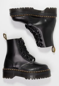 Bottes en cuir noir à lacets avec semelles épaisses rainurées et coutures jaunes, présentées vues de côté et de dessous sur fond blanc.