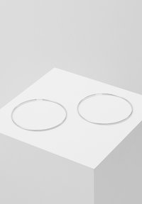 Deux boucles d'oreilles en métal argenté affichées sur un piédestal blanc. Les hoops sont fins, circulaires et au design minimaliste, avec une finition lisse.