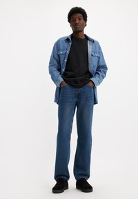 Chemise en denim bleu clair avec devant boutonné et poches, portée sur un T-shirt noir. Jean droit bleu foncé et chaussures noires.
