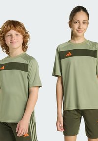 Dvě děti mají na sobě světle zelená sportovní trička s tmavě zelenými vodorovnými pruhy a oranžovými akcenty. Tkanina vypadá hladce a je lehká.