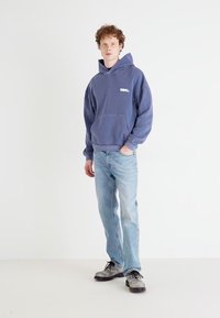 Blue cotton hoodie with a front pocket and a small logo, paired with light, faded jeans and gray shoes with black laces. Plain background.