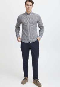 Button-up skjorte med smale vertikale svart og hvite striper, lange ermer og klassisk krage, parret med marineblå bukser og grå fottøy.
