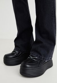 Svarta plattformsskor med texturerad yta, rund tå och kontrasterande grå snören, i kombination med mörk denimjeans som delvis syns.