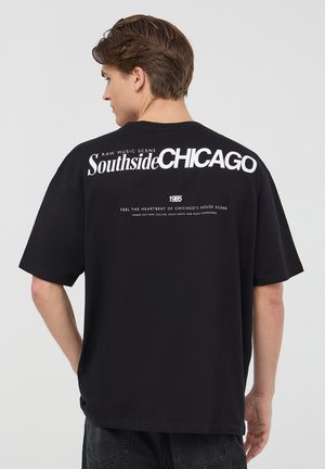 T-shirt in cotone nero con vestibilità comoda. Presenta una grande scritta bianca che dice "Southside CHICAGO" e dettagli testuali più piccoli sul retro.