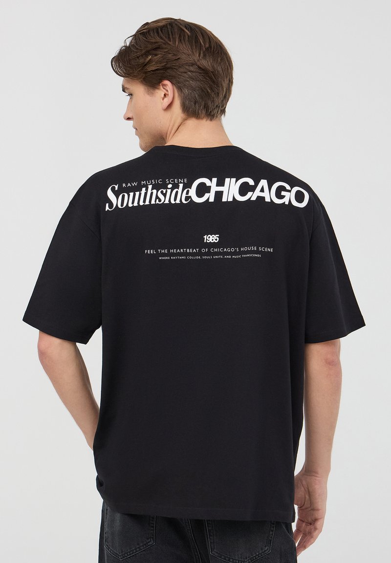 T-shirt in cotone nero con vestibilità comoda. Presenta una grande scritta bianca che dice "Southside CHICAGO" e dettagli testuali più piccoli sul retro.
