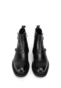 Botines de cuero negro con un acabado pulido, que incluyen cremalleras laterales, detalles de hebilla y un diseño de punta redonda. Suelas de goma gruesas y bajas.