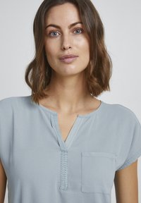 Lysblå kortærmet top med V-hals, frontlomme og subtil sy-detalje. Glat tekstur, afslappet pasform, casual design.