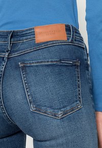Donkerblauwe spijkerbroek met achterzakken, voorzien van een tan leren taillebandlabel met de tekst "Marc O'Polo DENIM." Soepele textuur en aansluitend ontwerp.