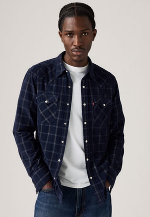 Navy geruite flanellen shirt met twee borstzakken, witte knopen en knoopsluiting, gedragen over een wit T-shirt, gecombineerd met denim jeans.