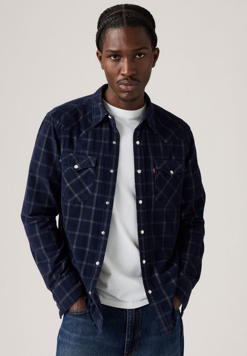 Navy geruite flanellen shirt met twee borstzakken, witte knopen en knoopsluiting, gedragen over een wit T-shirt, gecombineerd met denim jeans.
