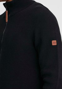 Sudadera negra con cremallera y cuello alto, hecha de un suave tejido de punto. Cuenta con un parche de cuero marrón con el logo en la manga izquierda.