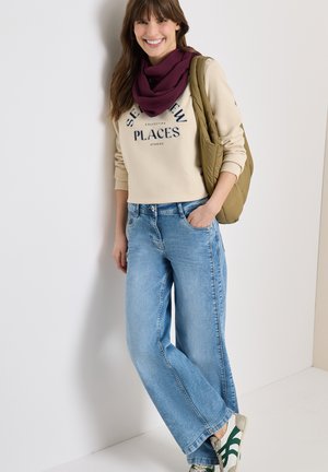 Vrouw lacht, draagt een beige sweatshirt met tekst, een bordeauxrode sjaal, lichtblauwe wijde jeans, witte sneakers en een groene schoudertas.