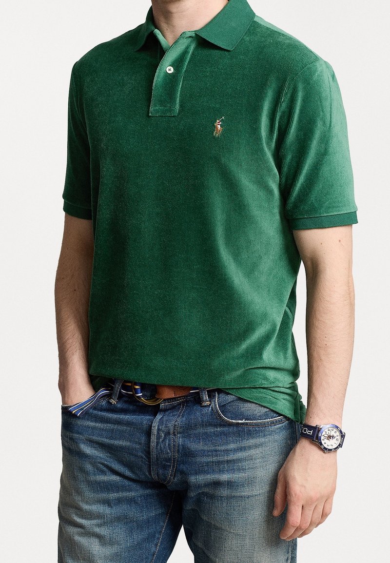Grøn, kordspun polo-shirt med krave og korte ærmer; har et lille broderet logo på brystet. Bæres med denim jeans.