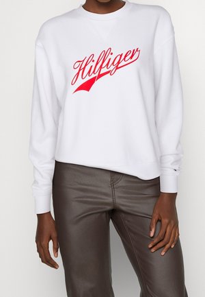 Persoon die een wit sweatshirt met lange mouwen draagt met een rood "Hilfiger" logo en donkerbruine leren broek, staand met ontspannen armen.