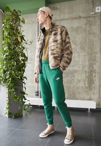 Fuzzy beige and brown camo jacket, green sweatpants with Nike logo, and tan high-top sneakers. Indoor setting with a plant.