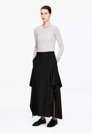 Femme portant un haut à manches longues gris clair, une jupe large asymétrique noire avec de grandes fentes, et des mocassins noirs, debout les mains dans les poches.