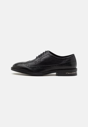 Schwarzer Brogue-Lederschuh mit Wingtip-Perforationen, Schnürverschluss und niedrigem Absatz vor einfachem weißem Hintergrund.