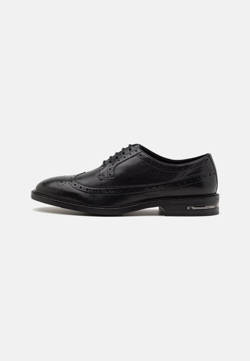 Chaussure brogue en cuir noir avec perforations wingtip, fermeture à lacets et faible talon sur un fond blanc uni.