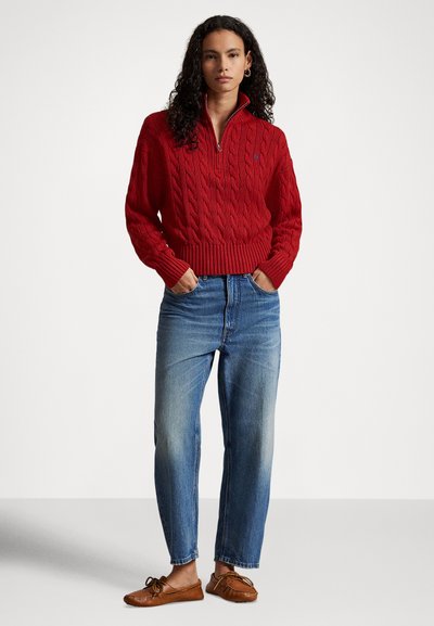 Γυναίκα με μπούκλες, που φοράει κόκκινο πουλόβερ με φερμουάρ στο λαιμό και πλεκτό σχέδιο, μπλε τζιν με τα χέρια στις τσέπες και καφέ μοκασίνια, στέκεται μπροστά σε λευκό φόντο.