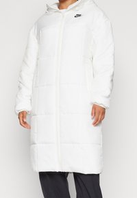 Vit puffjacka med huva, med en hel dragkedja fram, elastiska ärmslut och svart Nike-logotyp på övre vänstra bröstet.