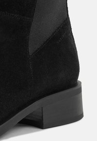 Juodos suede nėrinių aulenės su elastiniais šonų paneliais, apvaliu nosiu ir mažo blokinio kulniuko. Išskirtinės tekstūros kontrastai ir subtilus prekės ženklo detalė.