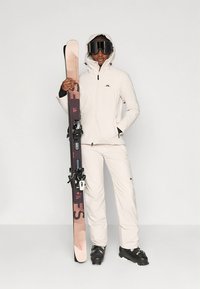 Biała kurtka narciarska i spodnie, czarne buty narciarskie, trzyma różowe i czarne narty. Strój posiada kaptur, zamki błyskawiczne i akcenty z logo.