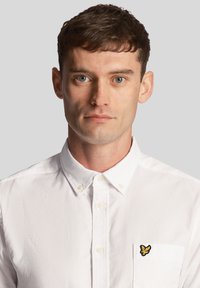 Camicia bianca con bottoni, caratterizzata da una finitura testurizzata. Presenta un piccolo logo giallo ricamato sulla tasca sinistra del petto. Design con colletto classico.