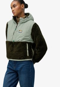 Veste à capuche doublée en polaire avec un dessus en nylon vert, des manches en fausse fourrure vert foncé et un ourlet côtelé ; présente un patch logo et des poches avant zippées.