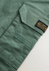 Section de pantalon cargo en tissu vert présentant une poche diagonale, des coutures texturées et une étiquette noire avec un texte doré. Fini de surface lisse.