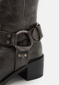 Czarny skórzany botek na kostkę z okrągłym metalowym pierścieniem i ozdobnym paskiem ze skóry. Wykończenie z widocznym przeszyciem i teksturą. Obcas typu słupek.