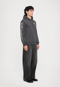 Male model standing, wearing dark gray EA7 Emporio Armani hoodie with logo on chest and sleeve, wide black jeans, and black shoes.