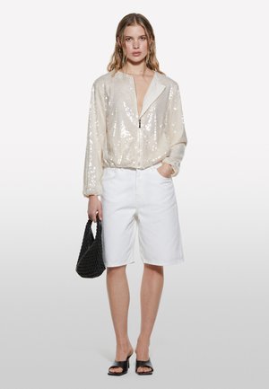 Giacca beige con paillettes e zip, pantaloncini di jeans bianchi, borsa intrecciata nera e sandali neri senza lacci. Presenta una texture liscia e una vestibilità comoda.