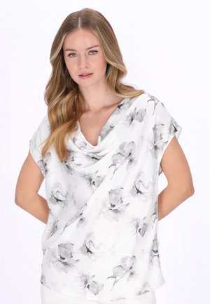 Femeie blondă cu părul ondulat, purtând o bluză albă cu mâneci scurte și modele florale gri, stând cu mâinile pe spate.