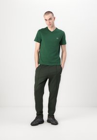 T-shirt in cotone verde con scollo a V, abbinato a pantaloni della tuta verde scuro con piping laterale e logo. Sneakers nere completano il look.