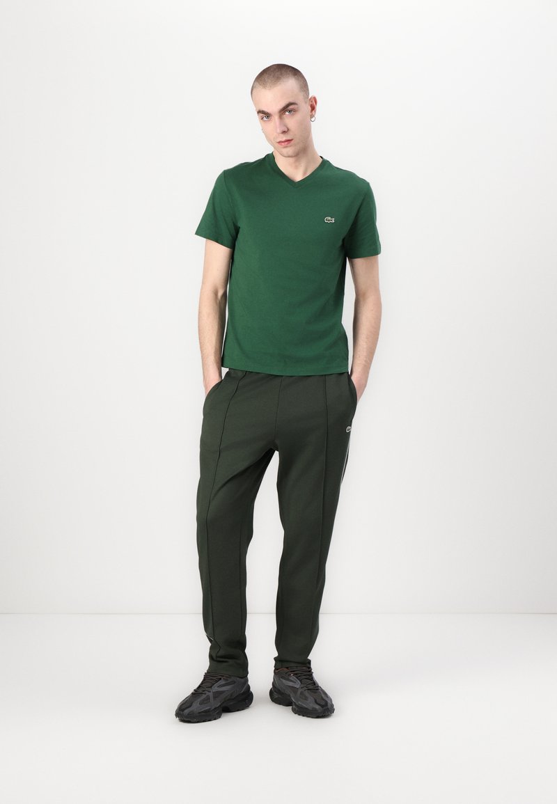 T-shirt en coton vert avec un col en V, associé à un pantalon de survêtement vert foncé avec des bandes latérales et un logo. Des baskets noires complètent le look.