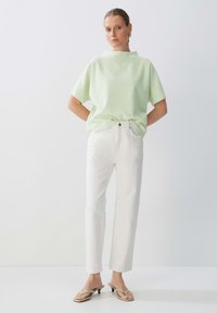Top verde chiaro strutturato con colletto alto, abbinato a jeans bianchi a gamba dritta e sandali con tacco beige. Design minimalista con una vestibilità rilassata.